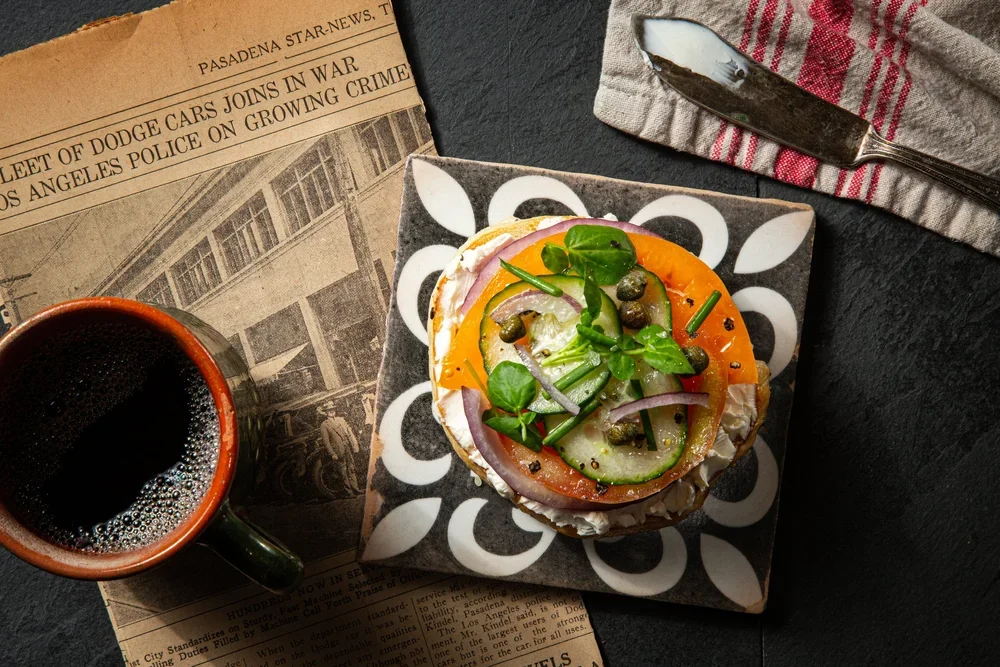 A plate of open-faced sandwich with sliced tomatoes, cucumbers, onions, capers, and greens on bread, alongside a cup of coffee on a dark surface with a newspaper and a fork on a striped cloth.