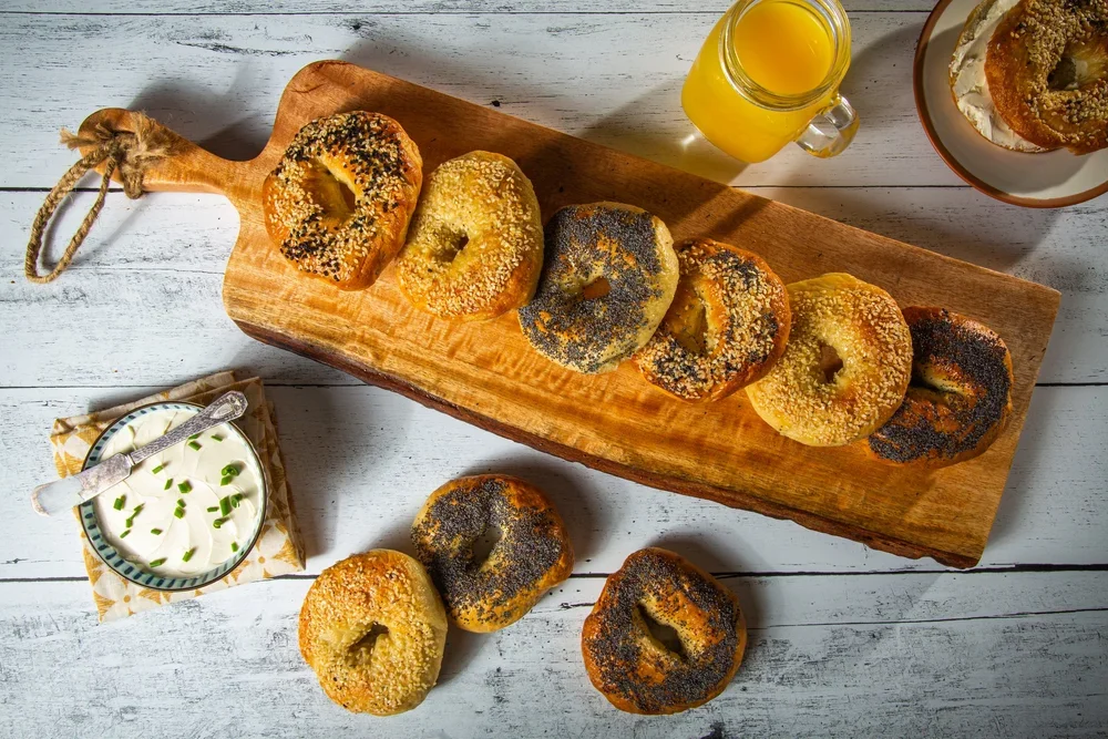 Assorted bagels with sesame and poppy seeds on a wooden serving board, accompanied by a bowl of cream cheese topped with chopped green onions, a glass of orange juice, and a bowl of a bagel with cream cheese on a white wooden table.