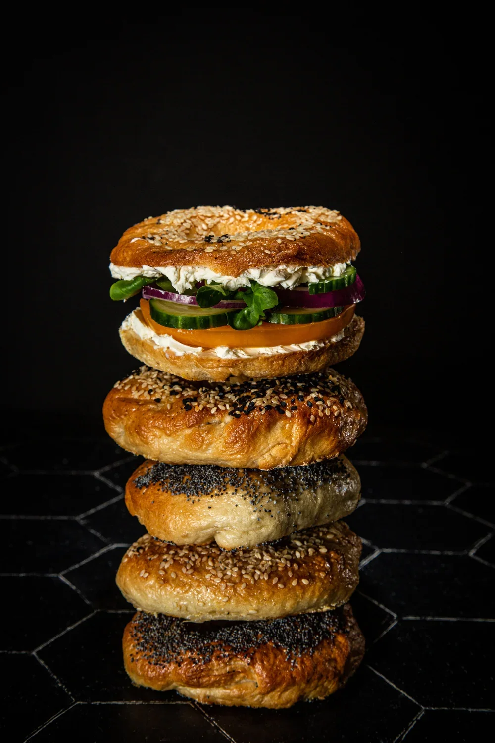 Stacked bagels with various toppings, including cream cheese, vegetables, and seeds on a black background.
