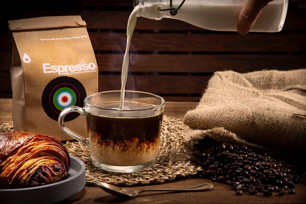 A glass cup of coffee with milk being poured into it, with a bag of espresso coffee, a chocolate croissant on a plate, scattered coffee beans, and a beige cloth napkin on a wooden table.