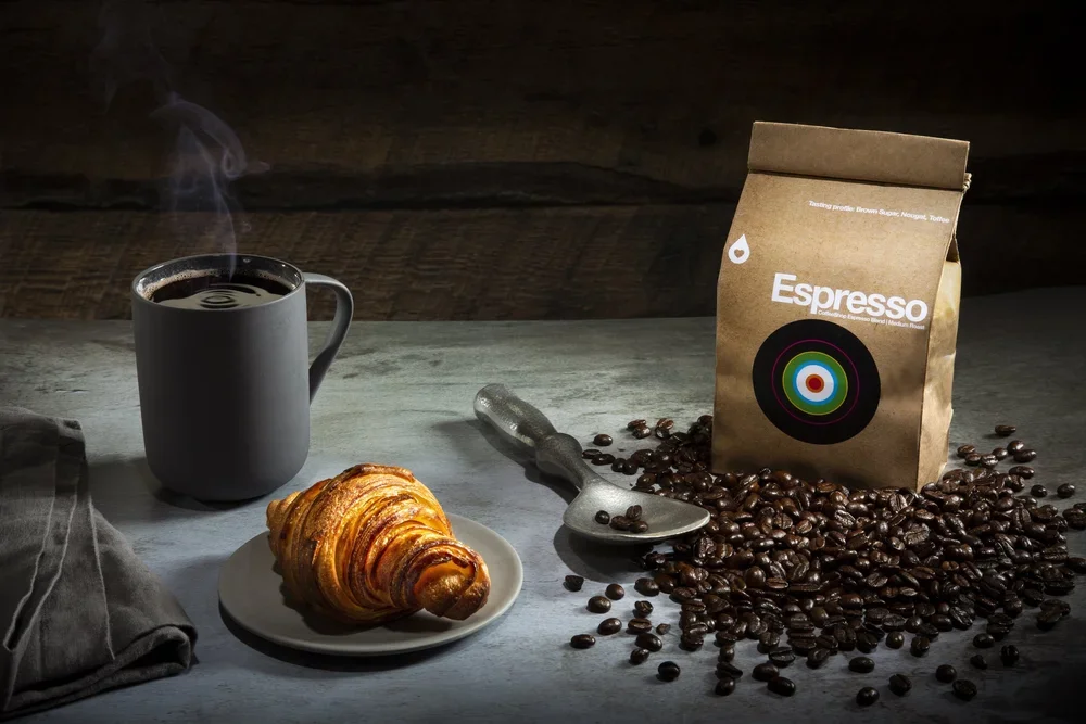 A steaming cup of black coffee, a croissant on a white plate, a bag of espresso coffee beans, and a spoon on a rustic table.