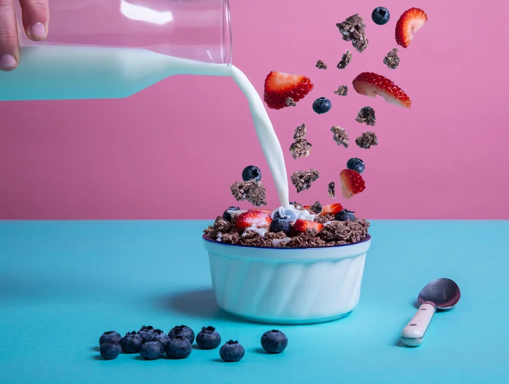 A bowl of cereal topped with strawberries, blueberries, and chocolate chunks, with milk being poured over it from a clear pitcher against a pink background, and a spoon on the blue surface.