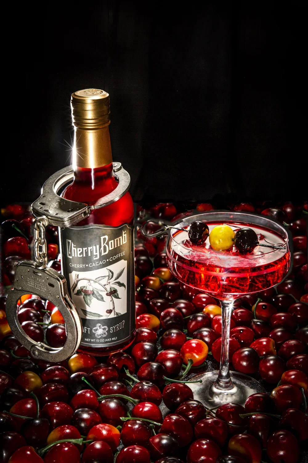 A bottle of Cherry Bomb cherry cacao coffee syrup with a gold cap, a handshake clasped with a pair of silver handcuffs, and a cocktail glass filled with a red drink garnished with cherries and a yellow cherry on top, all surrounded by fresh cherries 