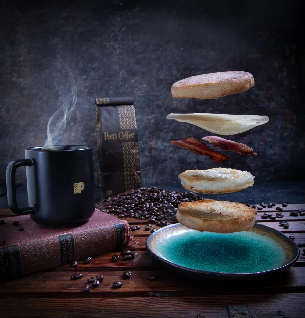 A black coffee mug with steam, a package of Peet's Coffee, a plate with floating breakfast items including toast, eggs, bacon, hash browns, and a plate with coffee beans, on a wooden table.
