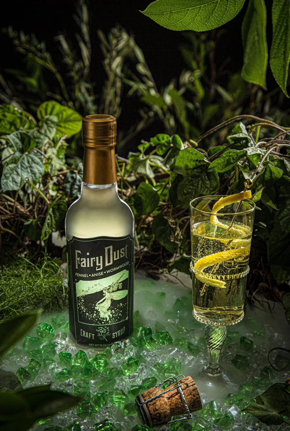 A bottle of Fairy Dust craft syrup labeled with herbs and spices, and a glass of drink with lemon twists and bubbles, surrounded by foliage and green and clear decorative gems.