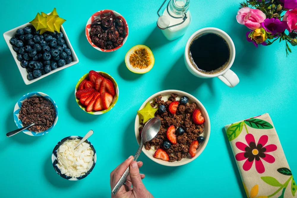 Breakfast scene with bowls of blueberries, strawberries, chocolate flakes, whipped cream, dates, a half passion fruit, a cup of black coffee, a brunch bowl with strawberries and blueberries, a bowl of dark chocolate shavings, a bottle of milk, a mug 