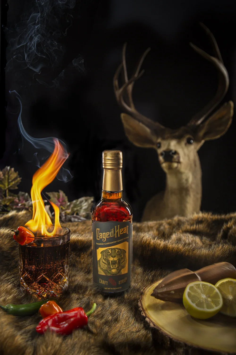 A bottle of CagedHeat craft syrup with a label featuring a fierce lion's head, surrounded by red and green chili peppers, lemon slices, a burning cocktail with a chili pepper garnish, a deer head with large antlers in the background, and a wooden pla