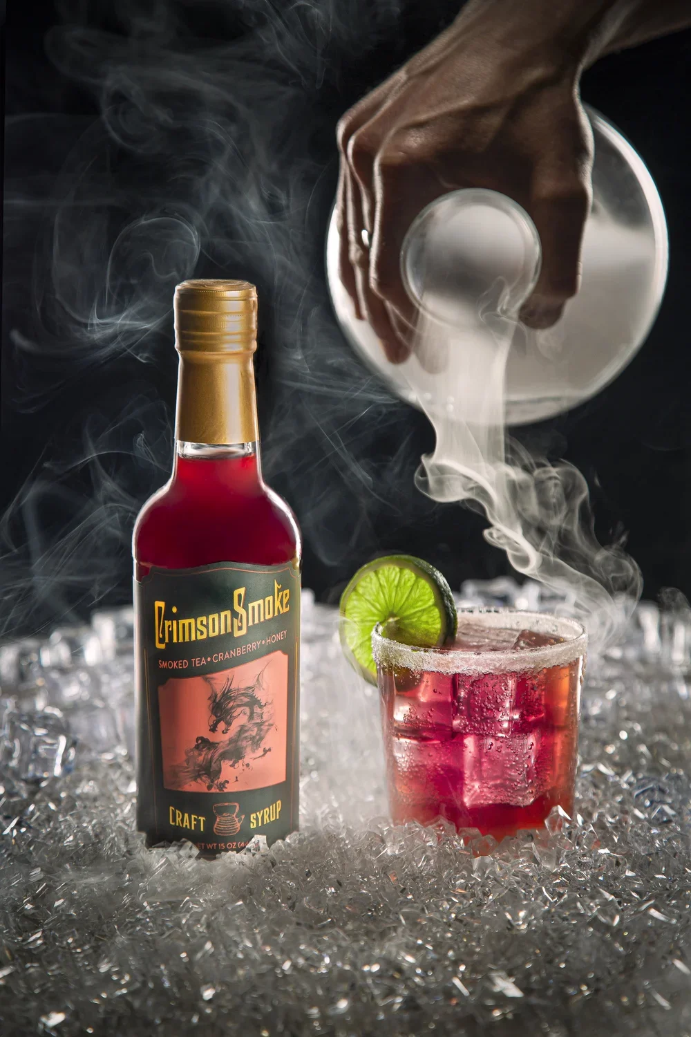 A hand pours liquid over a cocktail garnished with lime and cucumber on ice in a glass, with a bottle labeled 'Crimson Smoke' in the foreground and smoky vapor rising, set on crushed ice with a dark background.