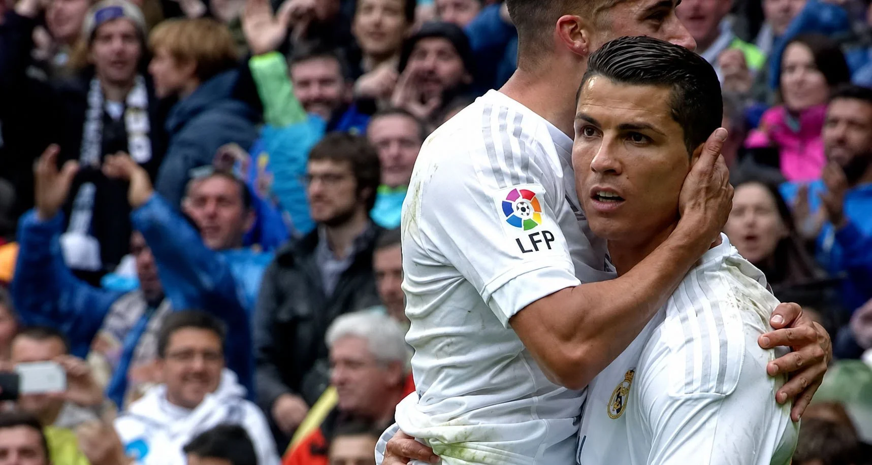 Two soccer players in white jerseys embrace on a field, surrounded by a crowd of spectators cheering.
