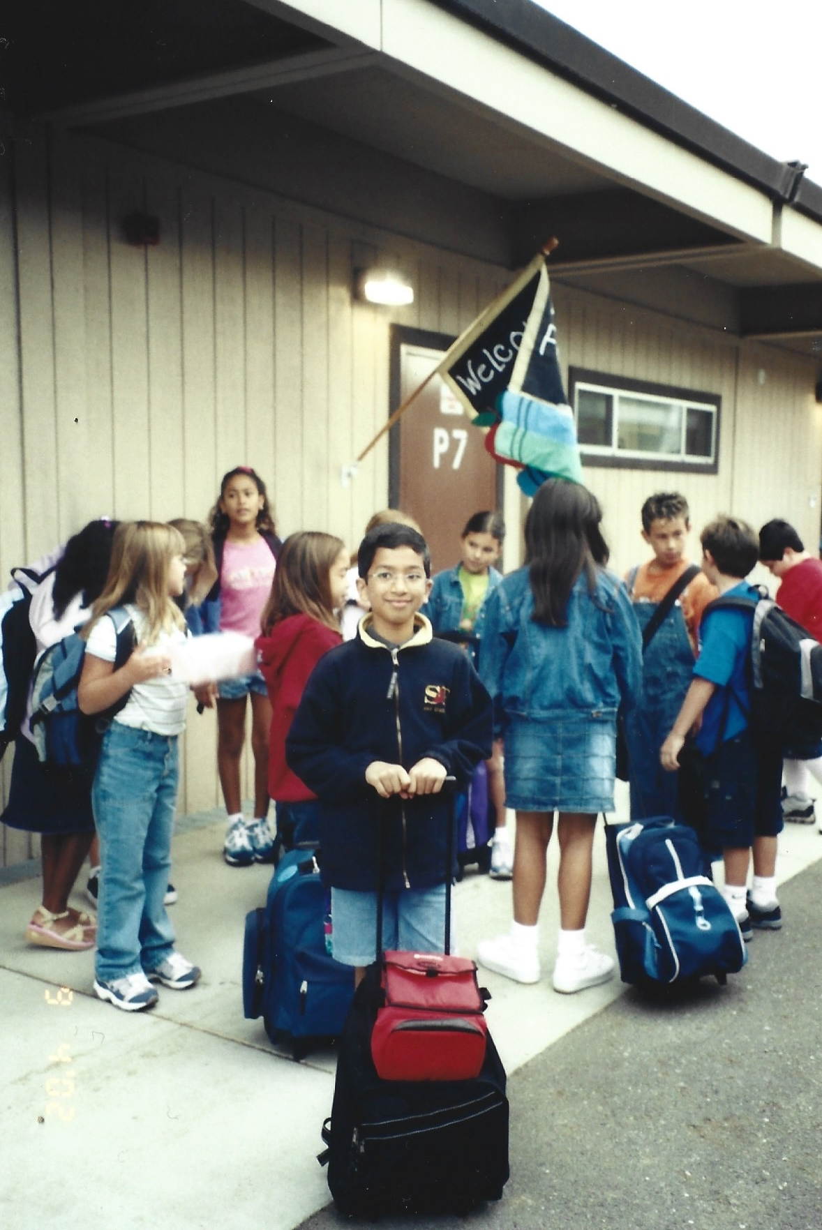 Group of children, likely students, standing outside a building with backpacks, holding luggage, and waiting for transportation or to enter the building, with a flag that says "Welcome" in the background.