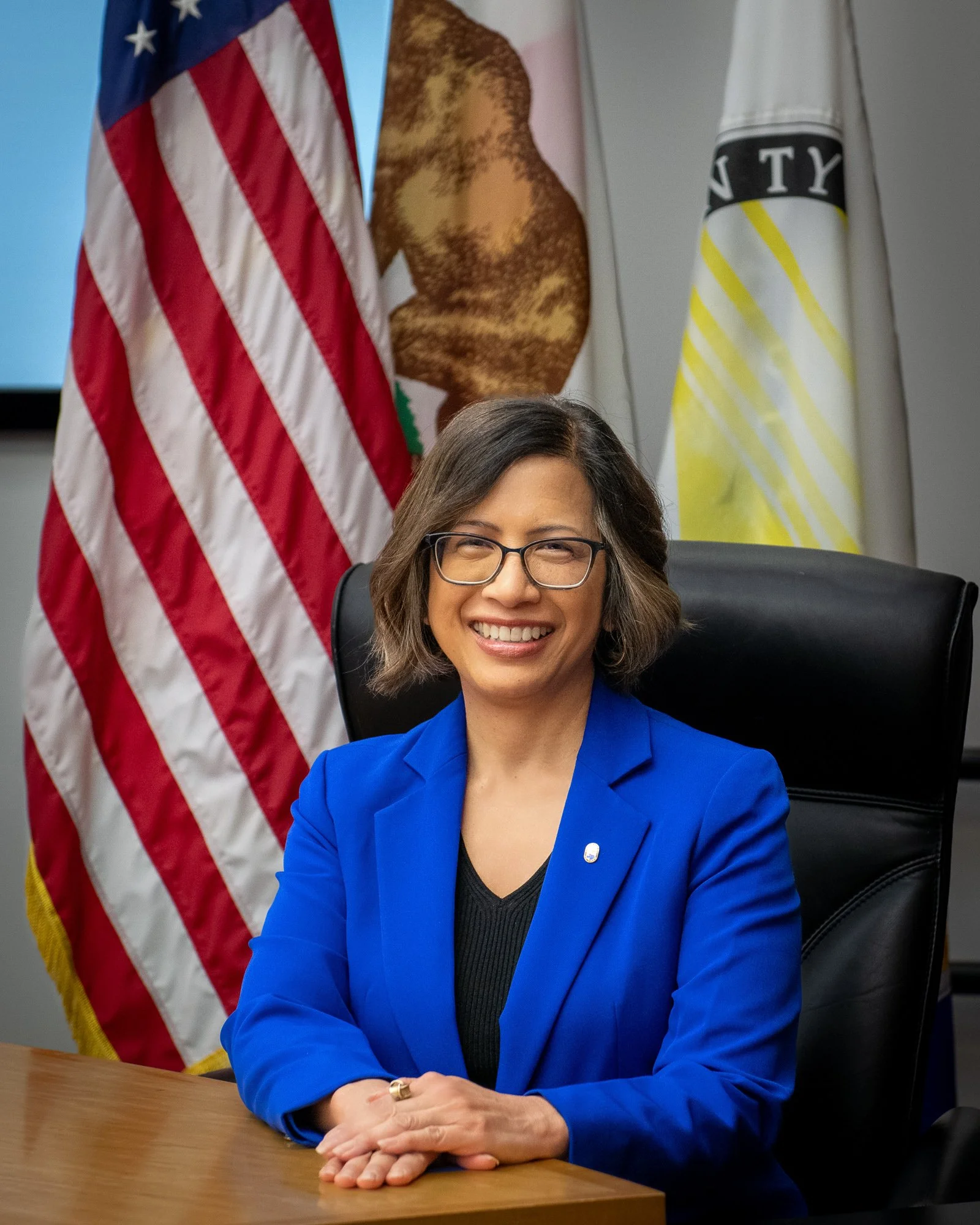 A woman with shoulder-length brown hair and glasses is sitting at a desk, smiling, wearing a blue blazer over a black top, with her hands clasped on the desk. Behind her, American and California state flags are displayed, with a portrait of a man in a suit visible between the flags.