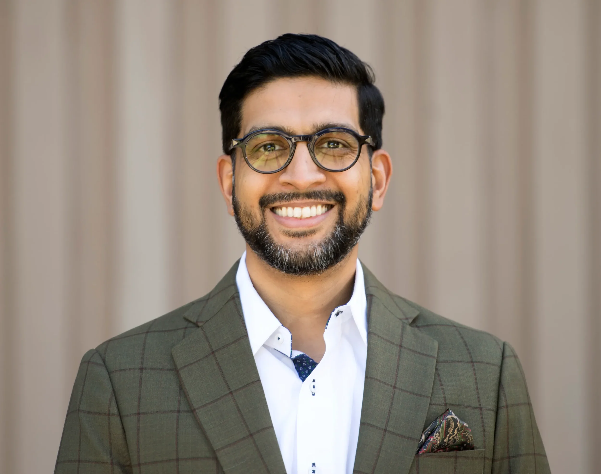 A man dressed in a green checkered suit, white shirt, and dark tie, smiling and wearing glasses, with a beige curtain background.