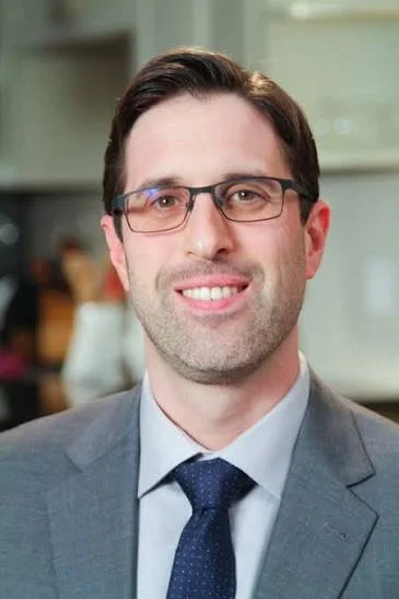 A man in a suit and tie with glasses, smiling in an indoor setting.