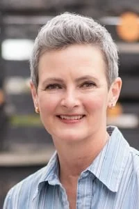 A woman outdoors smiling, with short gray hair, wearing a blue striped shirt.