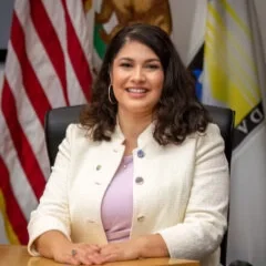Smiling woman sitting at a desk with American flags and governmental flags behind her.
