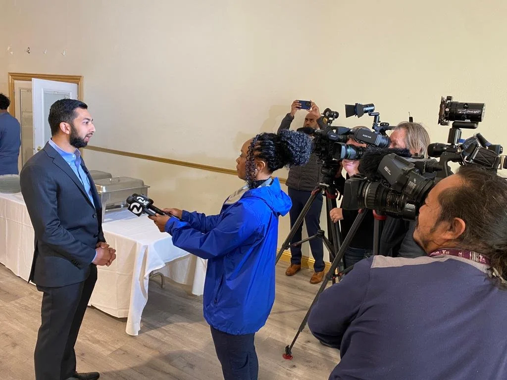 A man in a suit being interviewed by a female reporter with a microphone, surrounded by multiple videographers and photographers capturing the interview.