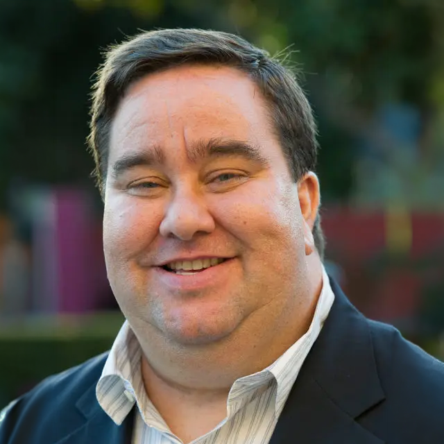 A smiling man in a dark suit jacket and white collared shirt standing outdoors with a blurred green and purple background.