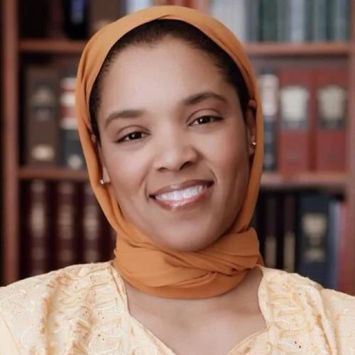 Close-up of smiling woman wearing an orange headscarf and cream-colored traditional clothing, with a bookshelf in the background.