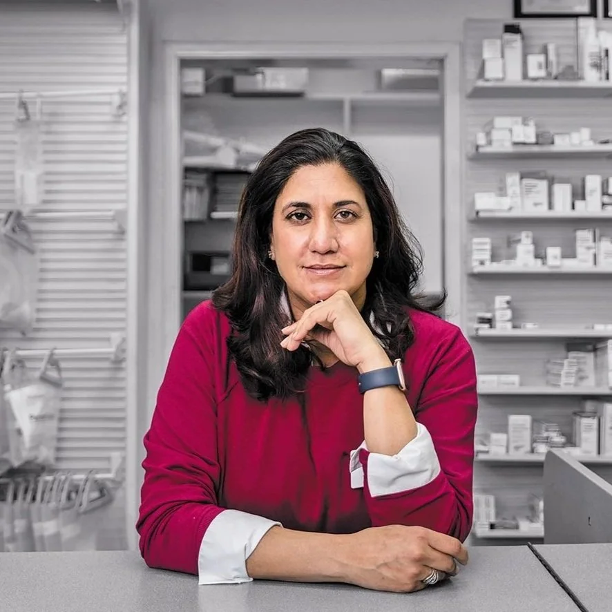 A woman with dark hair wearing a red top and a smartwatch, sitting at a counter in a pharmacy, with shelves of medication behind her.