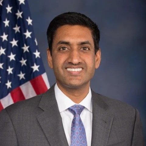 A professional man in a gray suit and tie, standing in front of an American flag backdrop.