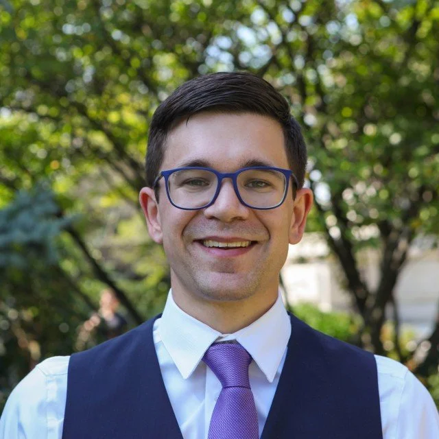 A young man with glasses smiling outdoors, wearing a white shirt, a purple tie, and a dark vest. Trees and greenery in the background.
