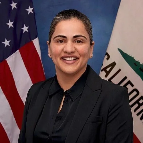 A woman with dark hair in a bun, smiling, wearing a black blazer and black shirt, standing in front of American and California flags.
