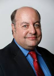 Close-up portrait of a middle-aged man with a light complexion, balding with hair on the sides, wearing a dark suit, blue shirt, and red tie, smiling slightly against a plain white background.