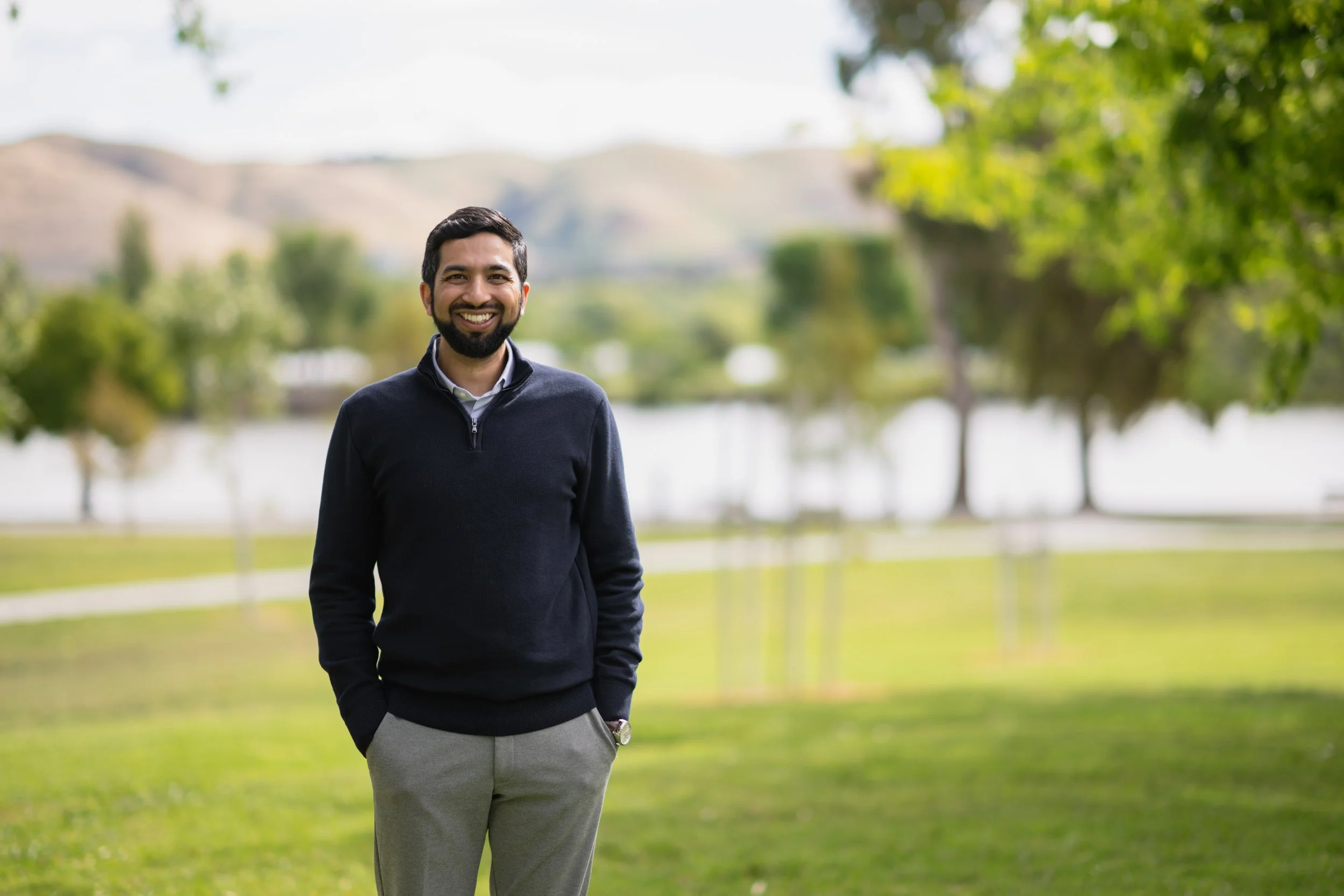 Smiling man with dark hair and beard standing outdoors in a park with green grass, trees, and a lake or river, wearing a dark sweater, light-colored pants, and a watch, with rolling hills in the background during daytime.
