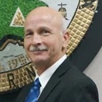 A man with a bald head and mustache in a suit and tie, standing in front of a colorful background with various symbols and text.