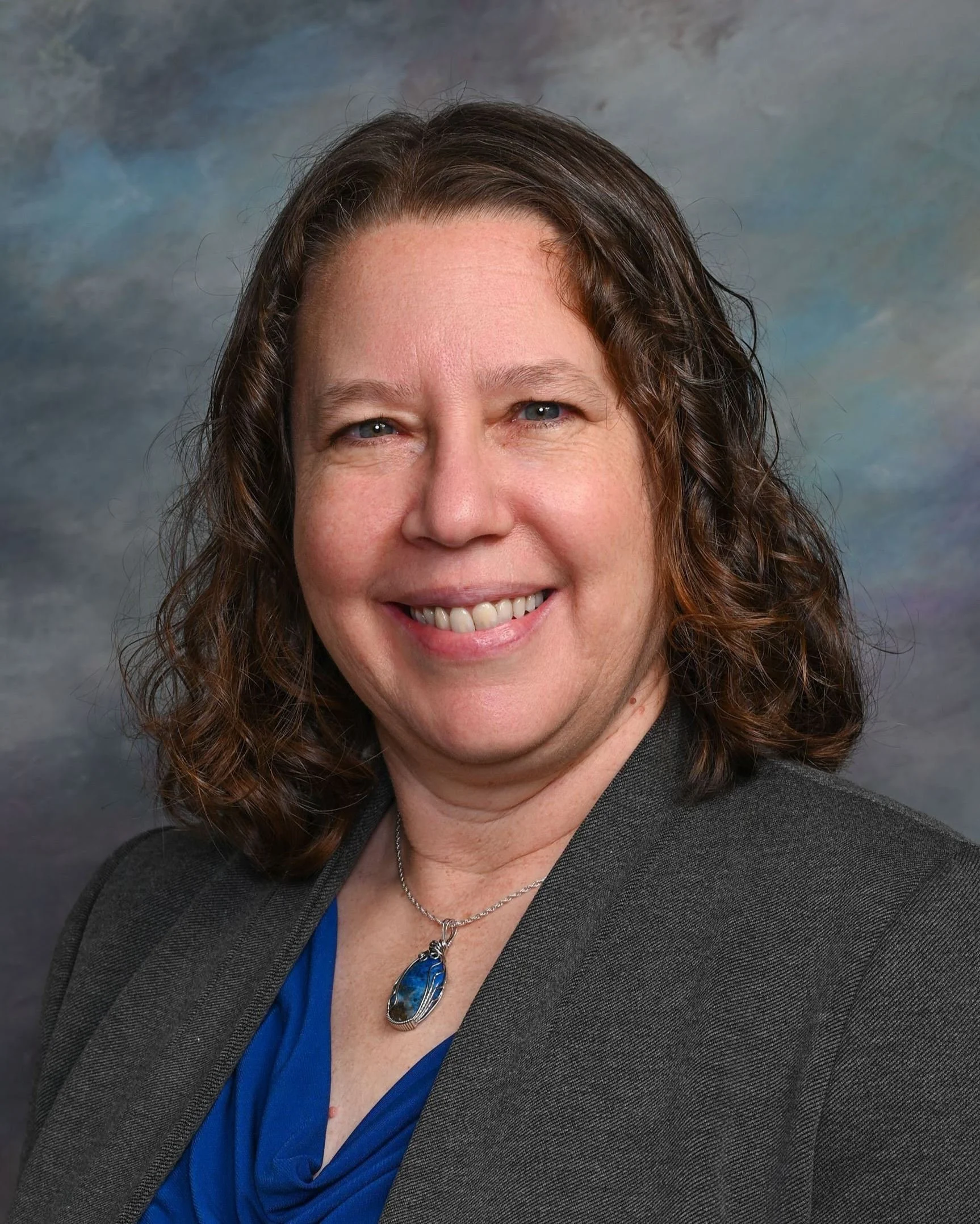 A woman with shoulder-length curly brown hair, wearing a gray blazer and a blue top, smiling and showing her teeth, with a pendant necklace featuring a blue gemstone, standing against a cloudy sky background.