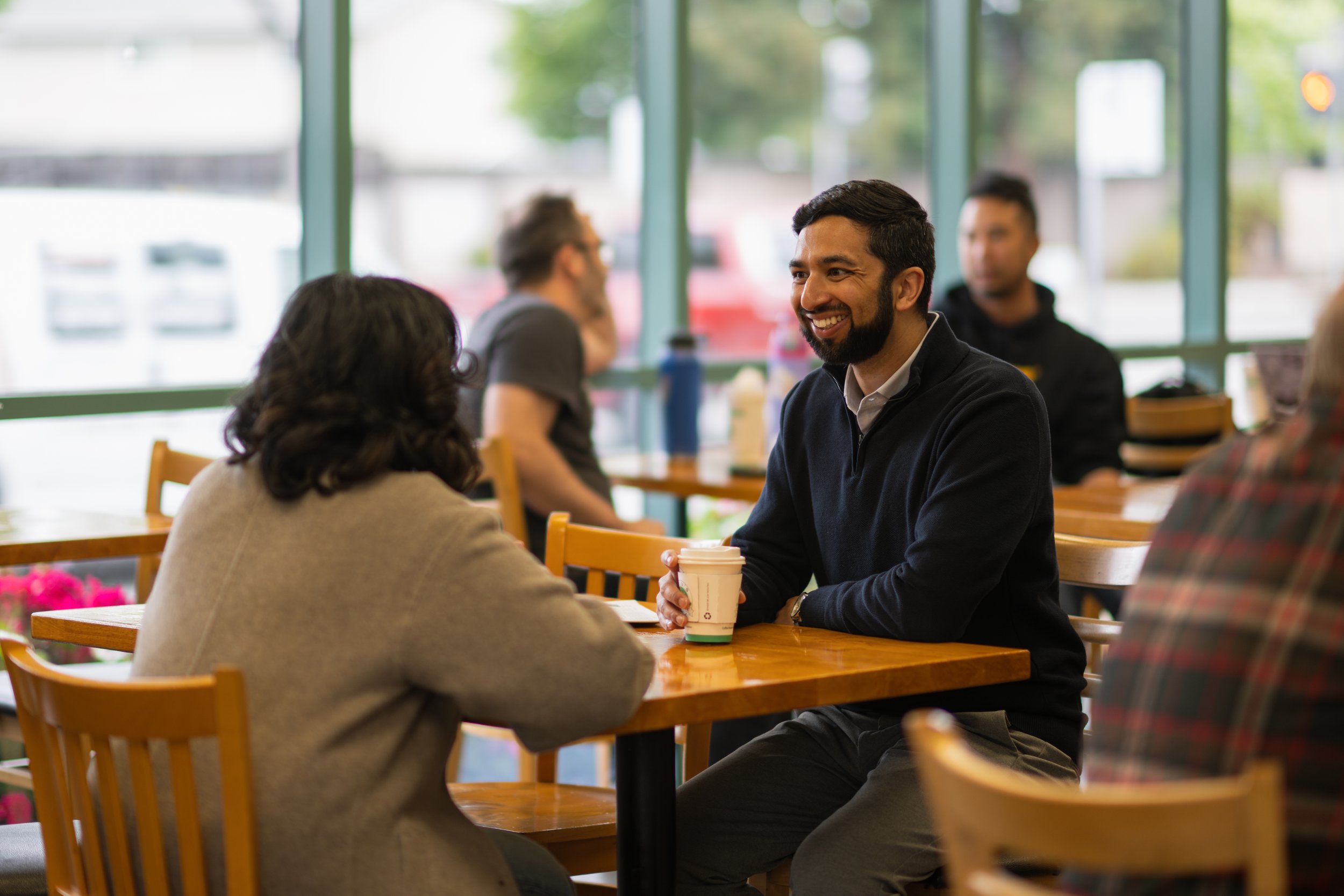 A man with a beard and dark hair smiling while sitting at a wooden table in a coffee shop, talking to a woman with dark hair. Other people are visible in the background near large windows with green frames, and daylight is coming through the windows.