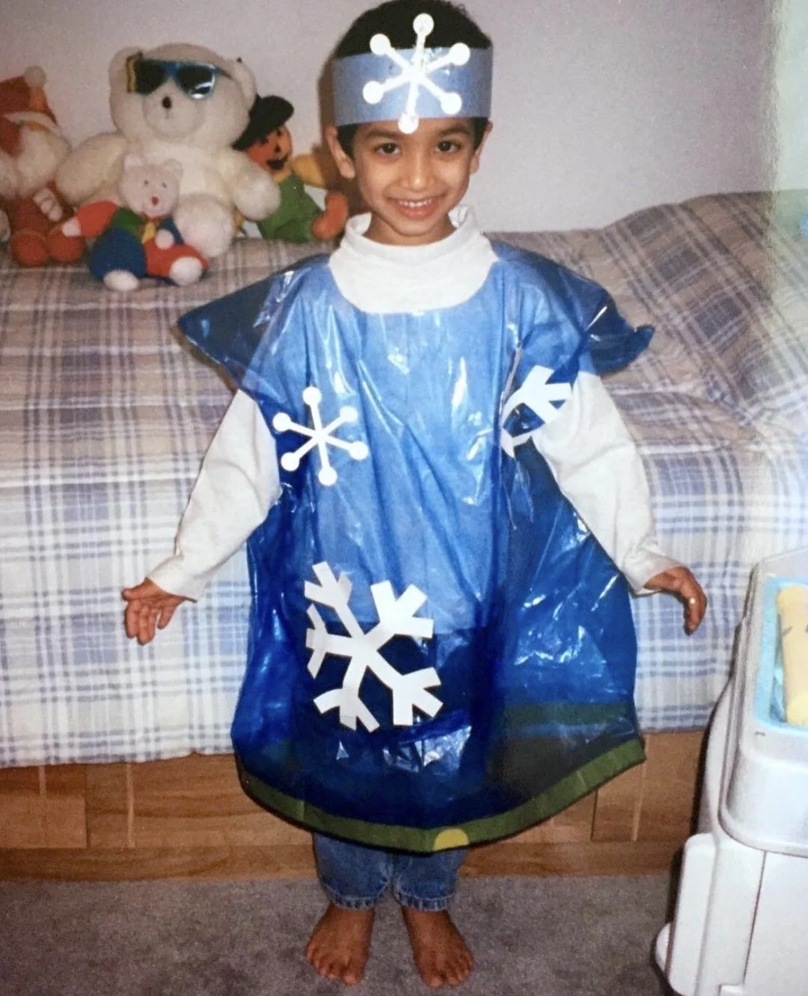 A young boy dressed in a homemade snowflake costume, featuring blue plastic with white snowflake cutouts, standing in a living room with a couch and stuffed animals in the background.