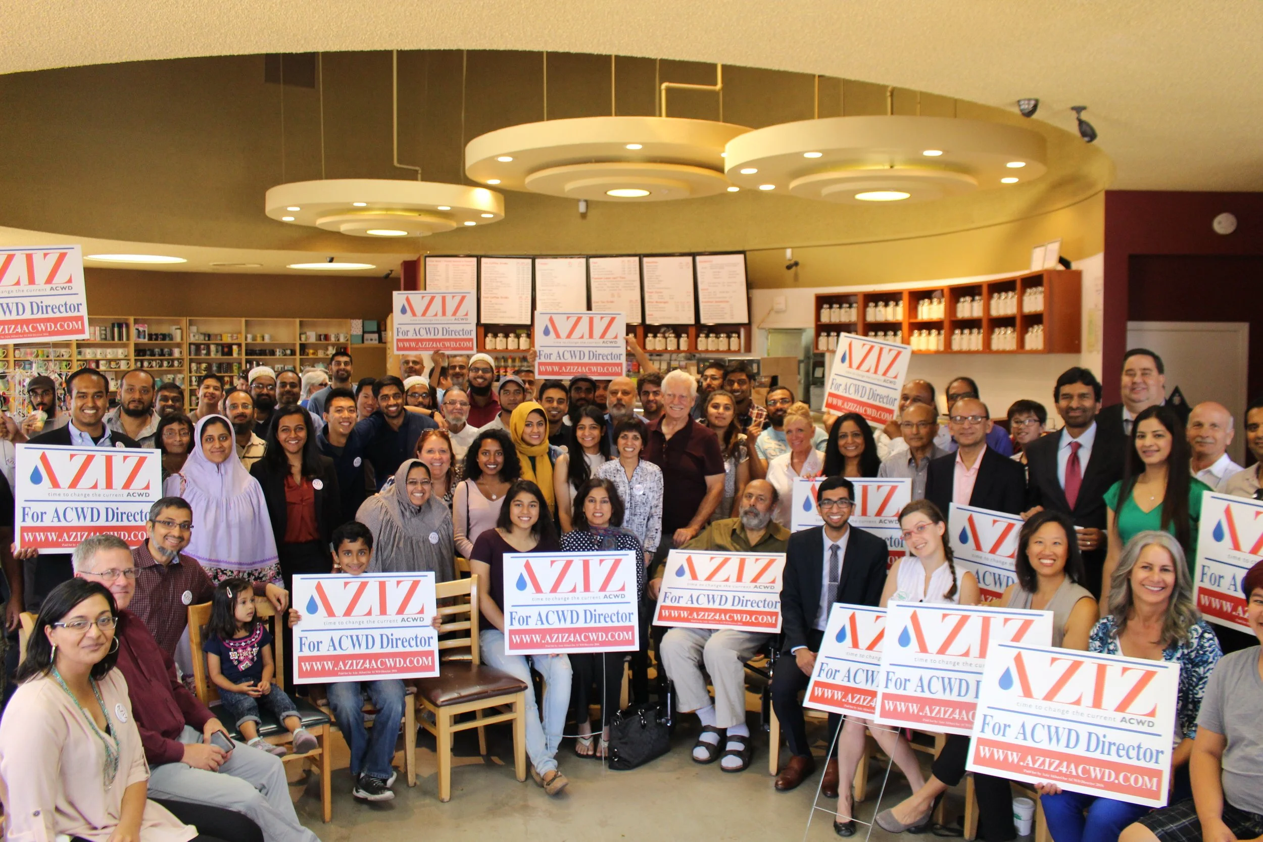A large group of diverse people gathered indoors, holding signs that read "AZIZ for ACWD Director" with the website www.aziz4acwd.com, smiling and posing for a group photo in a store or community space.