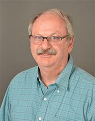 Portrait of a man with glasses, gray hair, and a mustache, wearing a light blue checkered shirt, smiling against a gray background.