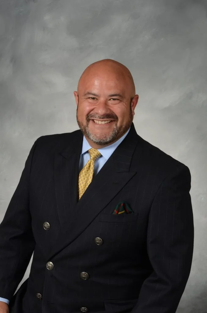 A smiling man with a bald head and a beard, wearing a dark pinstripe suit, a light blue shirt, a yellow patterned tie, and a pocket square, standing against a neutral background.