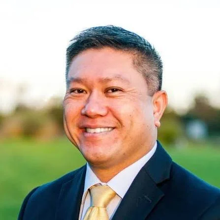 Portrait of a smiling man in a suit outdoors with a blurred green background.