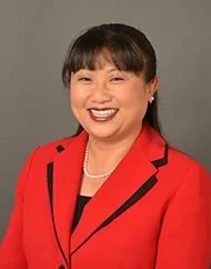 A woman wearing a red blazer and black shirt, smiling against a gray background.