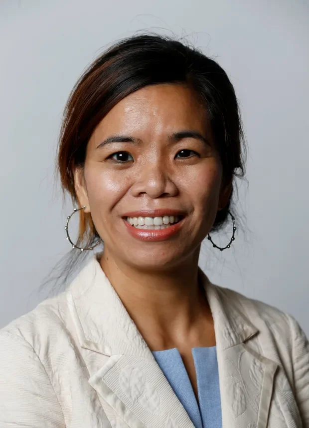 Portrait of a woman smiling, wearing earrings and a light-colored blazer, against a plain background.