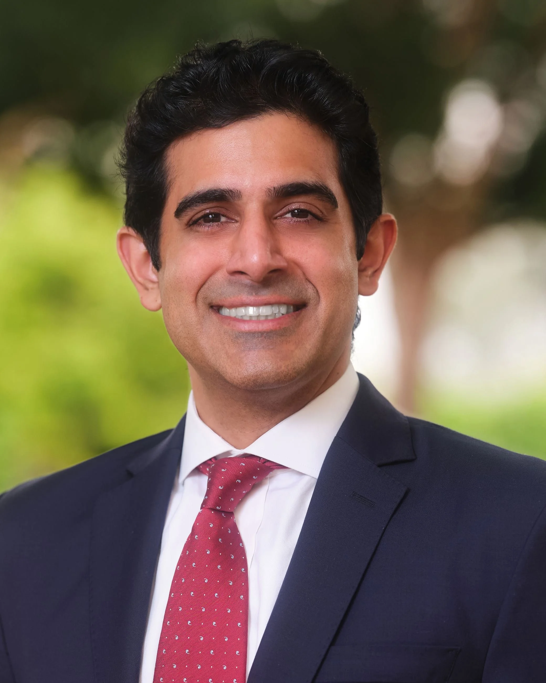 Close-up of a smiling man wearing a dark suit, white shirt, and red tie with white polka dots, outdoors with blurred green background.