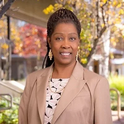 A woman with styled hair and earrings standing outdoors with autumn trees in the background.
