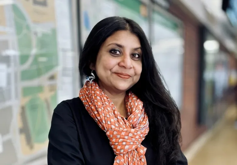 A woman with long black hair, wearing a black top and orange patterned scarf, standing outdoors near a blurred background of a map and glass storefronts.