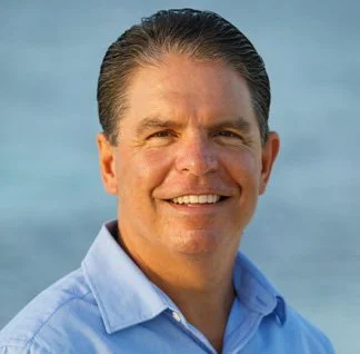 A smiling man with dark hair, wearing a blue collared shirt, outdoors with a blue sky in the background.