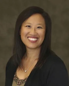 Portrait of a smiling woman with dark hair and a necklace, wearing a black top, against a dark background.