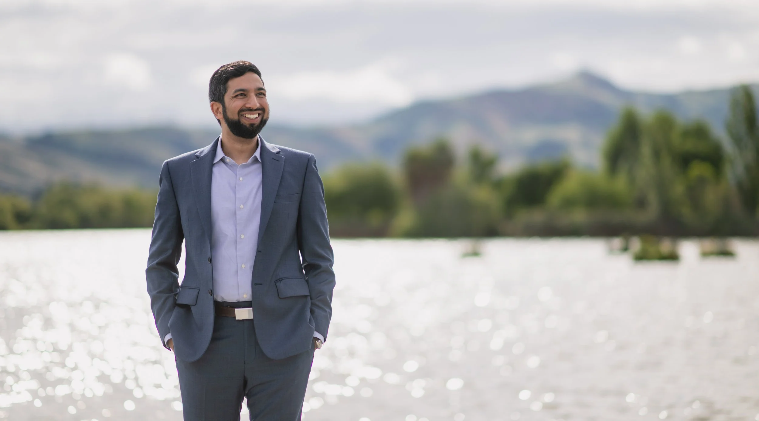 A man in a gray suit with a white shirt smiling and walking outdoors by a body of water with green trees and mountains in the background.