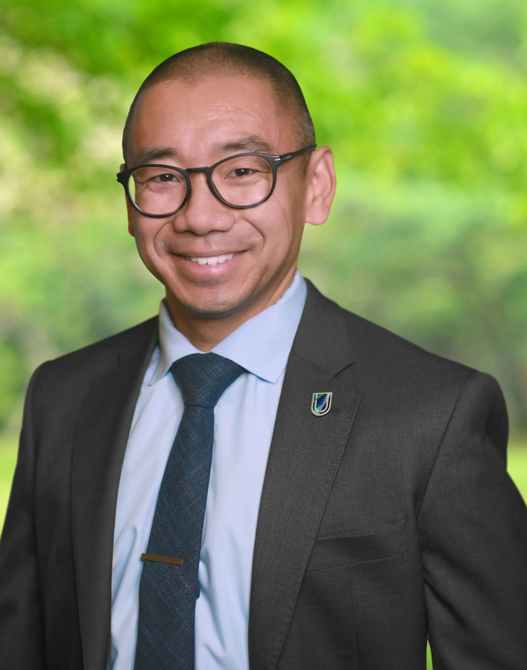Close-up of a smiling man wearing glasses, a dark suit, white shirt, and dark tie, with a blurred green outdoor background.