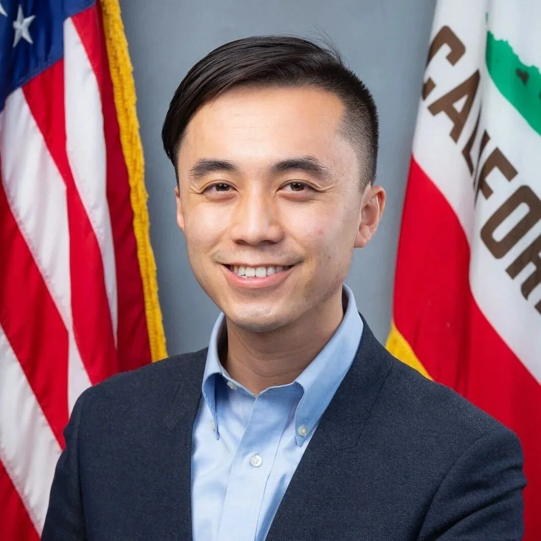 Portrait of a smiling man in a suit with American and California flags in the background.