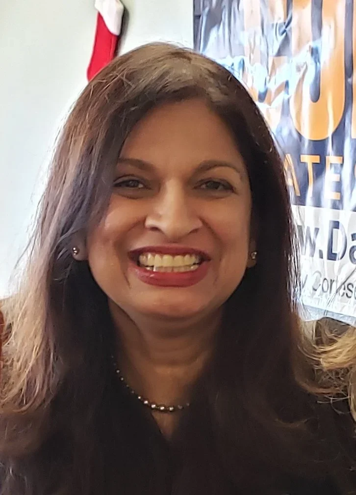 Smiling woman with long brown hair wearing earrings and a pearl necklace, indoors with a flag and red and white Christmas stocking in the background.