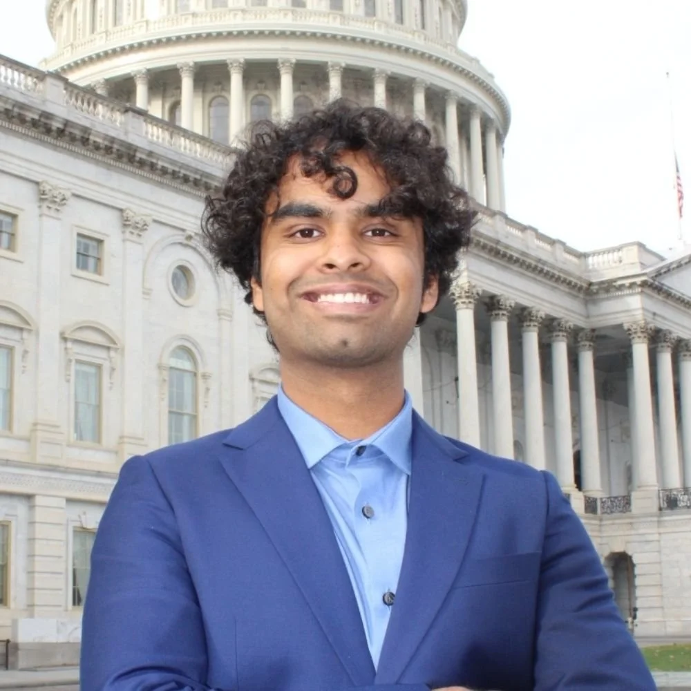 A man in a blue suit standing in front of the U.S. Capitol building, smiling with arms crossed.