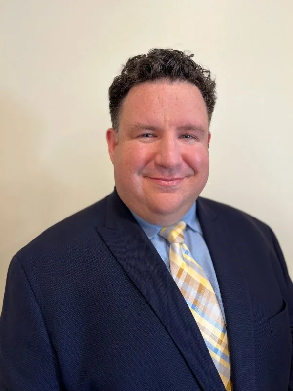 A man in a dark blue suit with a light blue dress shirt and a yellow, white, and light blue plaid tie, standing against a plain, light-colored background with a slight smile.