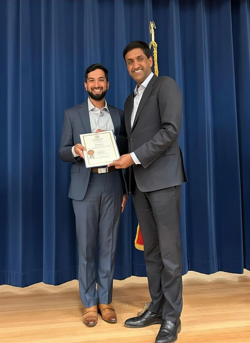 Two men in suits standing on a stage with a blue curtain behind them. One man is holding a certificate with a gold seal, and they are smiling.